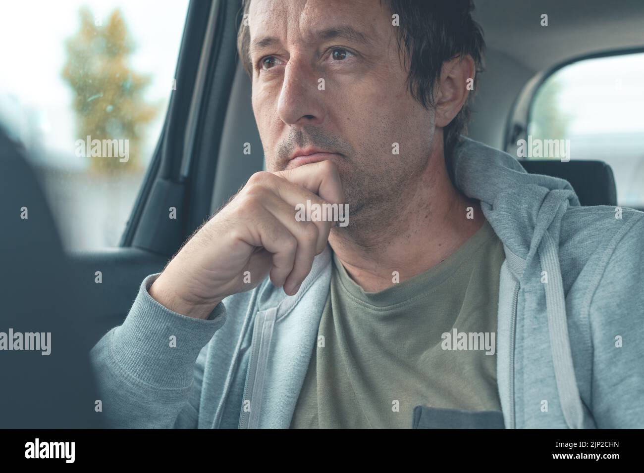 Hombre contemplativo con sudadera gris claro con capucha sentado en el asiento trasero del coche y pensando, enfoque selectivo Foto de stock