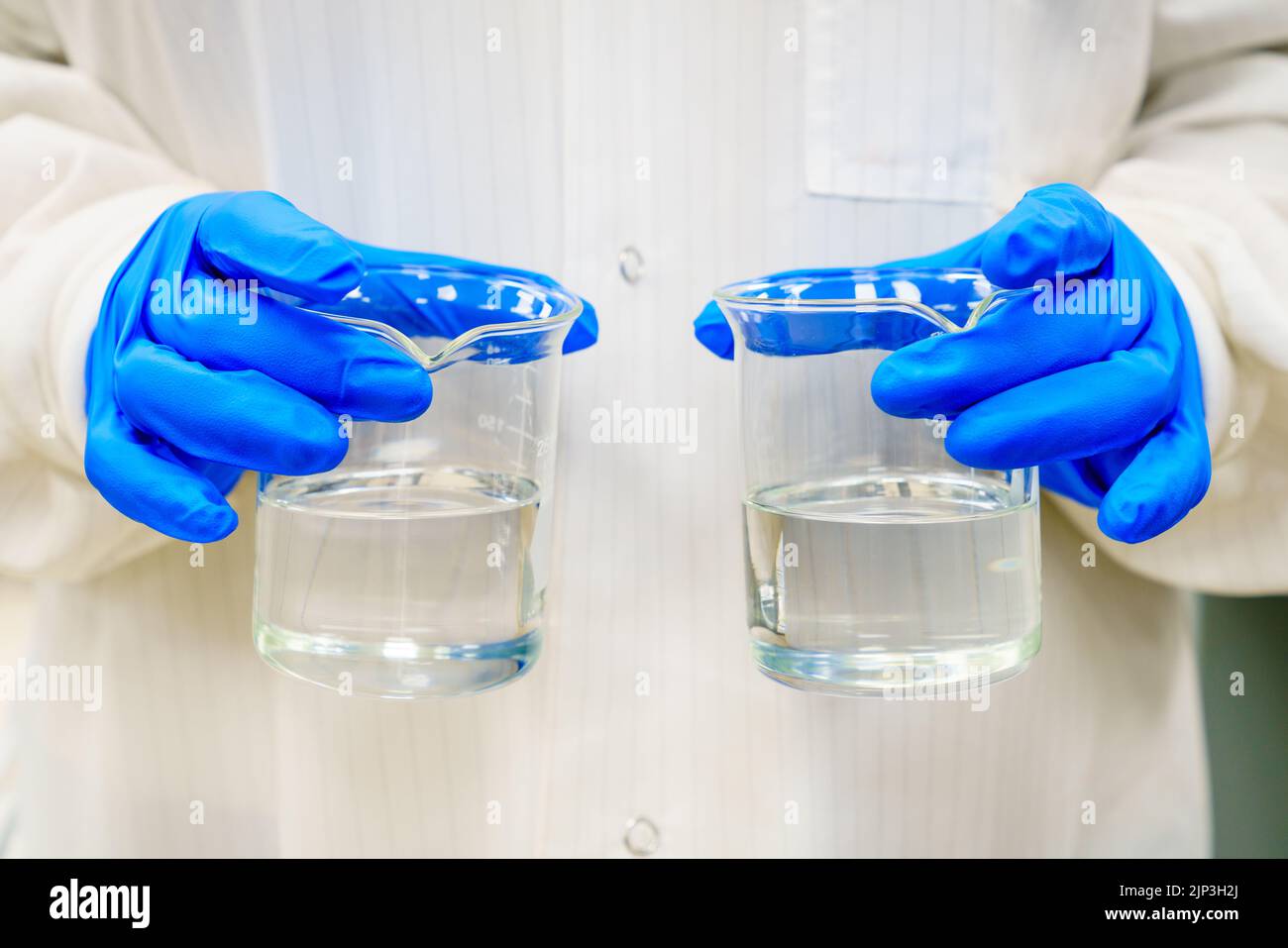 Una persona en una bata de laboratorio está sosteniendo dos vasos de precipitados con líquidos claros Foto de stock
