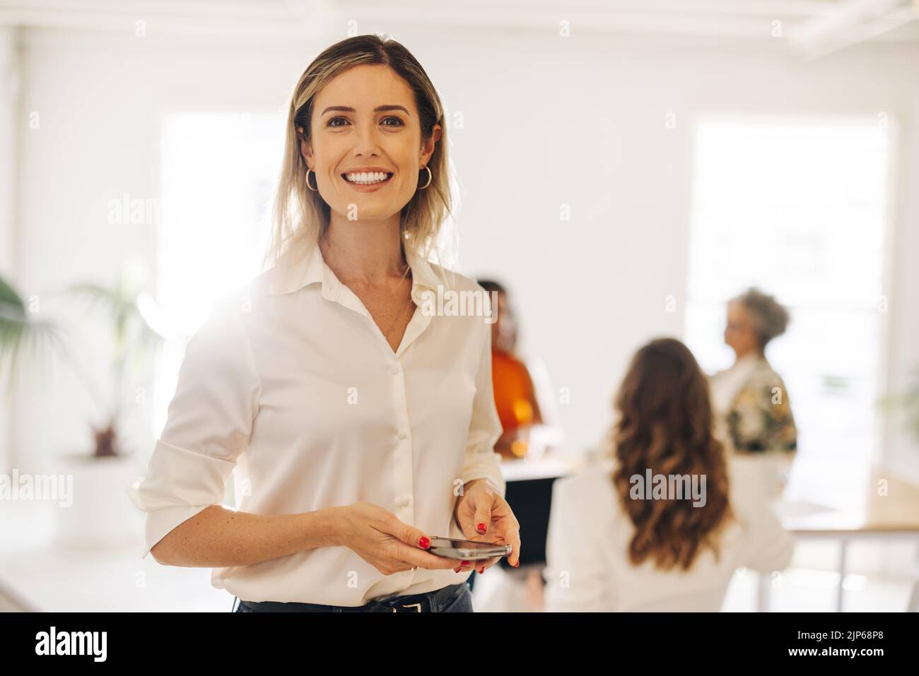 Alegre joven empresaria sonriendo a la cámara mientras sostiene un smartphone en una sala de reuniones. Feliz mujer joven de negocios trabajando en un todo-mujer de Foto de stock