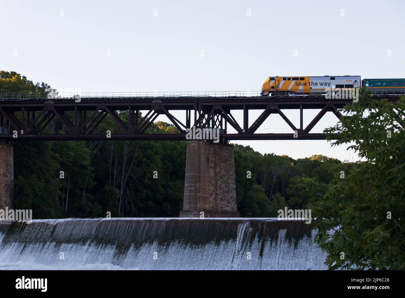 Un tren DE pasajeros VIA Rail Canada se ve pasando por un puente sobre un río, con una cascada. Foto de stock