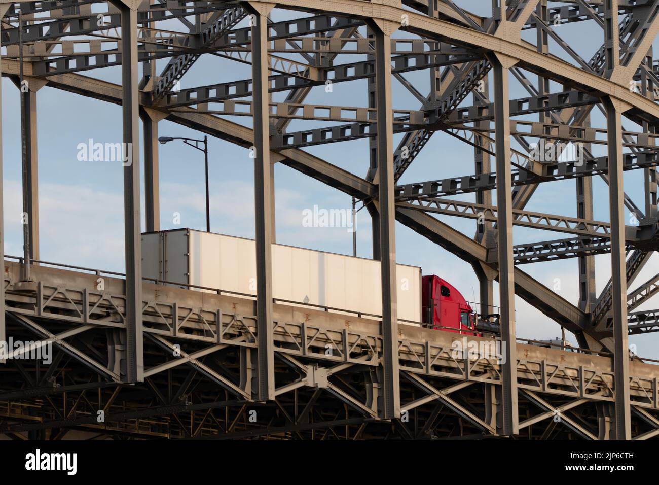 Se ve un camión de transporte rojo con un remolque blanco viajando a través de un puente sobre la armadura. Foto de stock