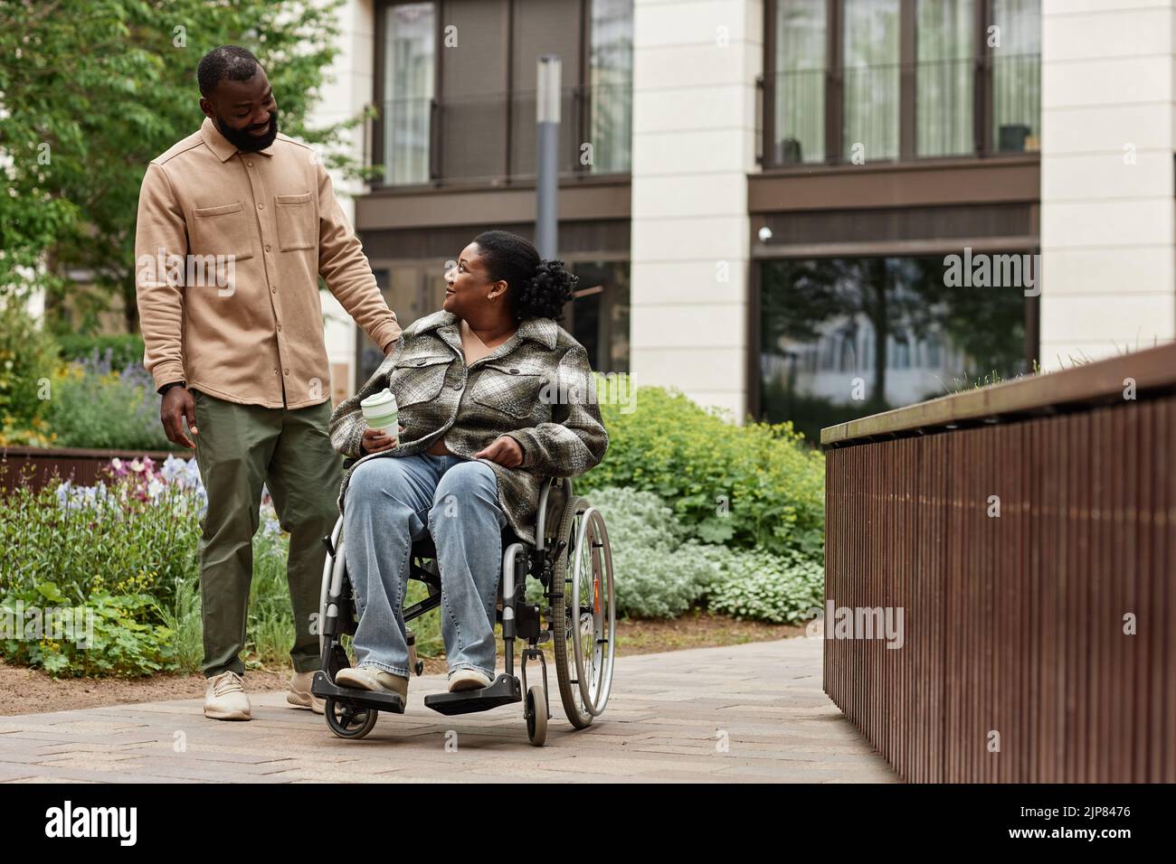Retrato completo de una pareja negra feliz con una mujer joven en silla de ruedas disfrutando caminar en el jardín de la ciudad juntos y mirándose con amor, c Foto de stock