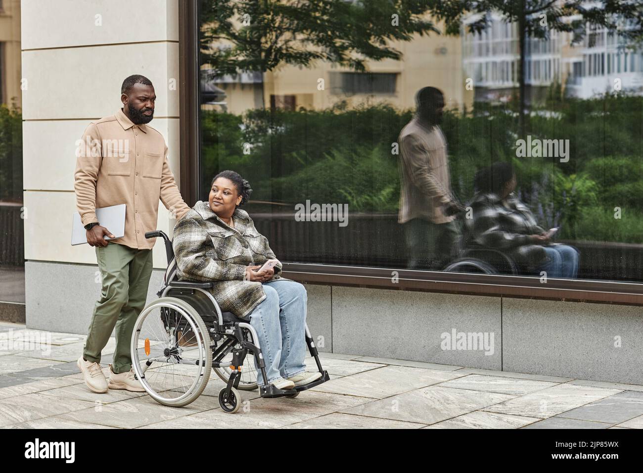 Retrato completo de la pareja negra moderna con la pareja en silla de ruedas al aire libre en el entorno de la ciudad, espacio de copia Foto de stock