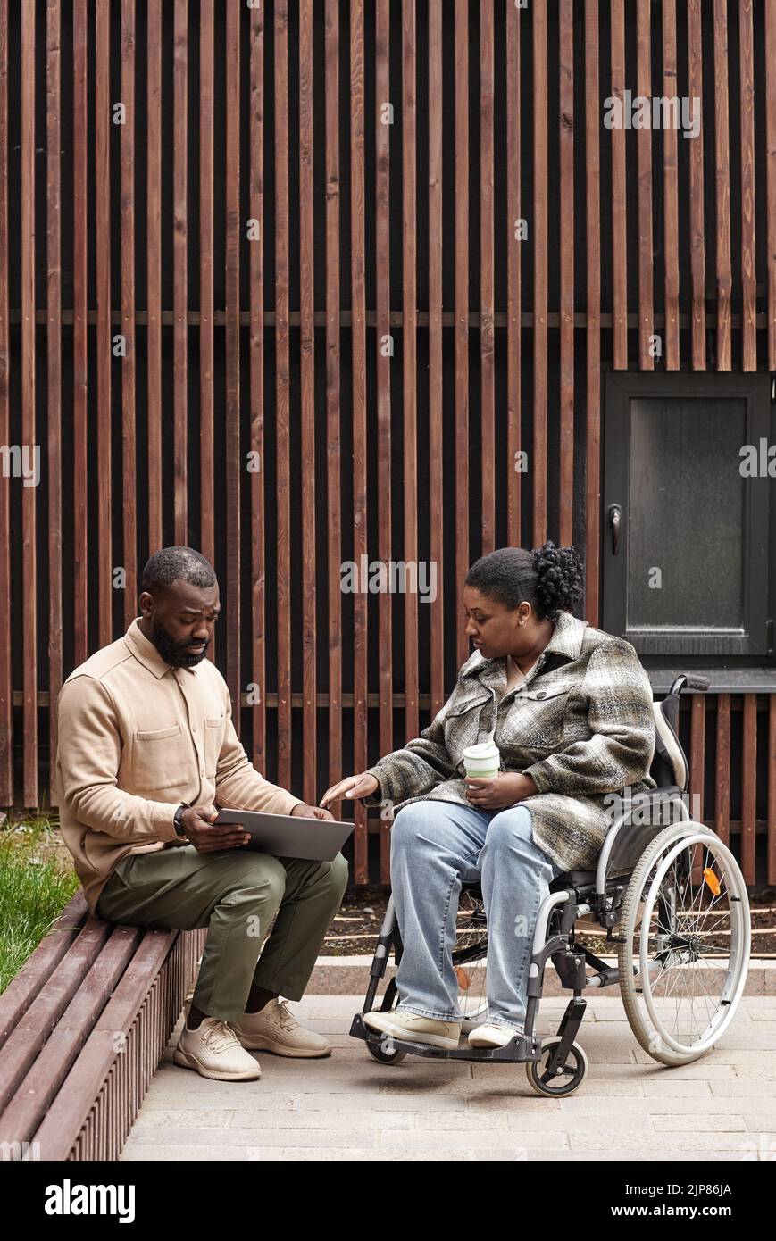 Plano vertical completo de una mujer joven en silla de ruedas hablando con un amigo o colega al aire libre en el parque de la ciudad, espacio de copia Foto de stock