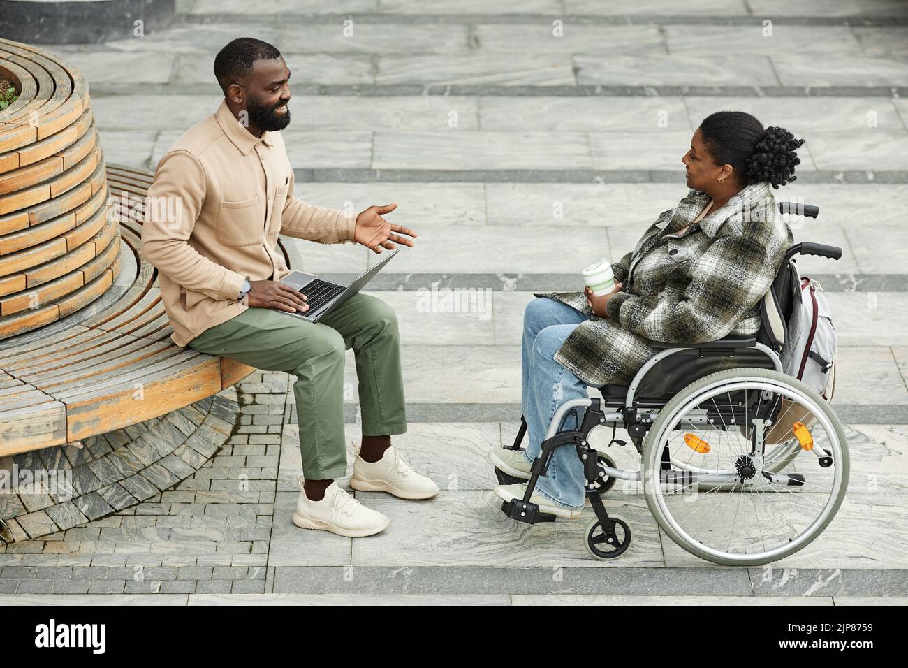 Fotografía completa de una pareja negra adulta con una mujer en silla de ruedas, charlando al aire libre en un entorno urbano y disfrutando del café Foto de stock