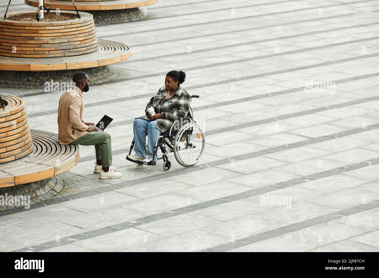 Gráfico gran angular de la pareja de adultos con mujer negra en silla de ruedas chateando al aire libre en la ciudad entorno con piso de baldosas, espacio de copia Foto de stock
