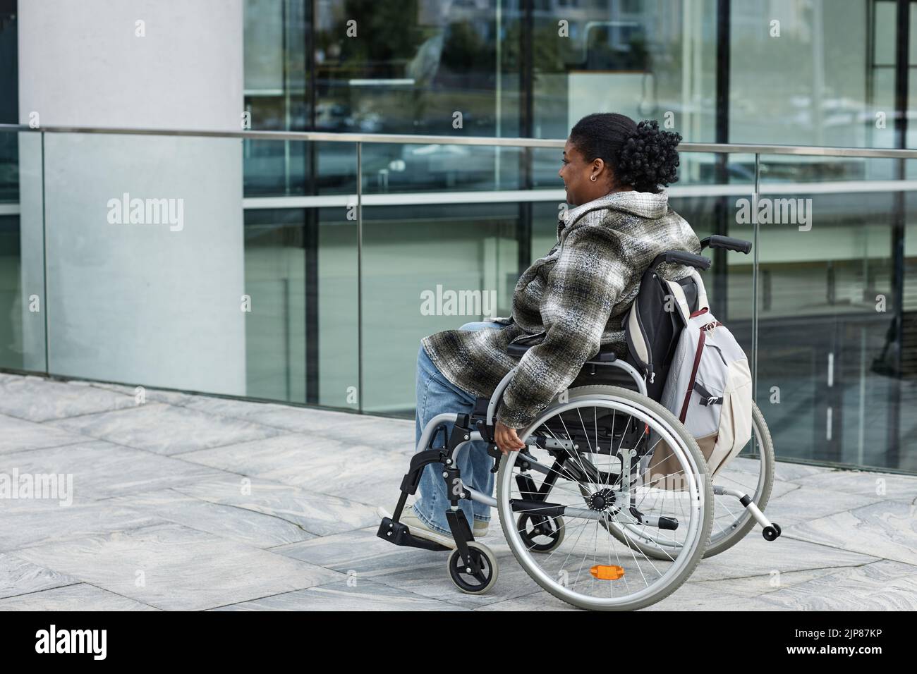 Fotografía completa de una mujer negra adulta en silla de ruedas desplazándose en un entorno urbano, espacio para copias Foto de stock