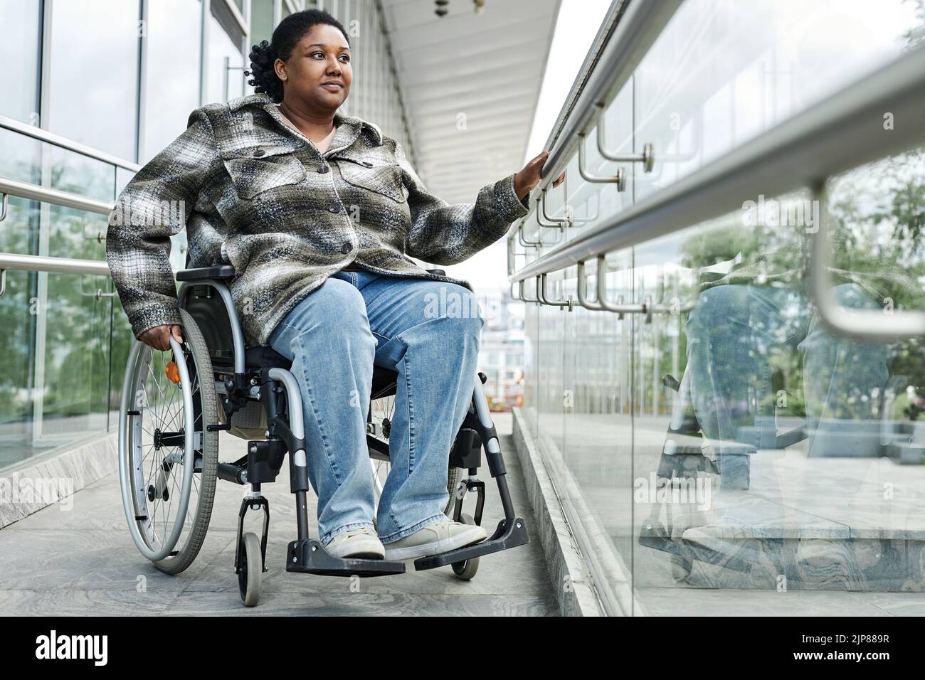 Retrato completo de una mujer negra sonriente en silla de ruedas bajando por rampa en la ciudad, función de accesibilidad urbana, espacio de copia Foto de stock