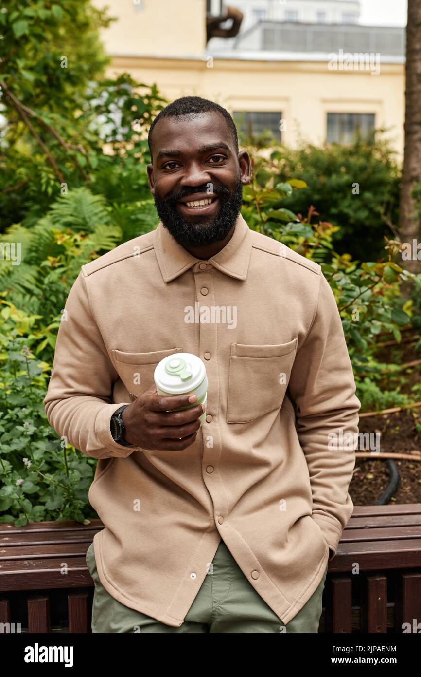 Retrato vertical de cintura arriba de un hombre negro sonriente sosteniendo una taza de café en el jardín de la ciudad y mirando la cámara Foto de stock