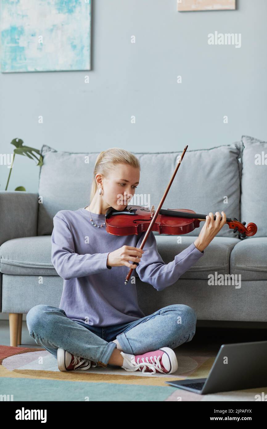 Retrato vertical completo de una joven rubia tocando violín en casa mientras estaba sentada en el suelo y viendo una clase de música en línea Foto de stock