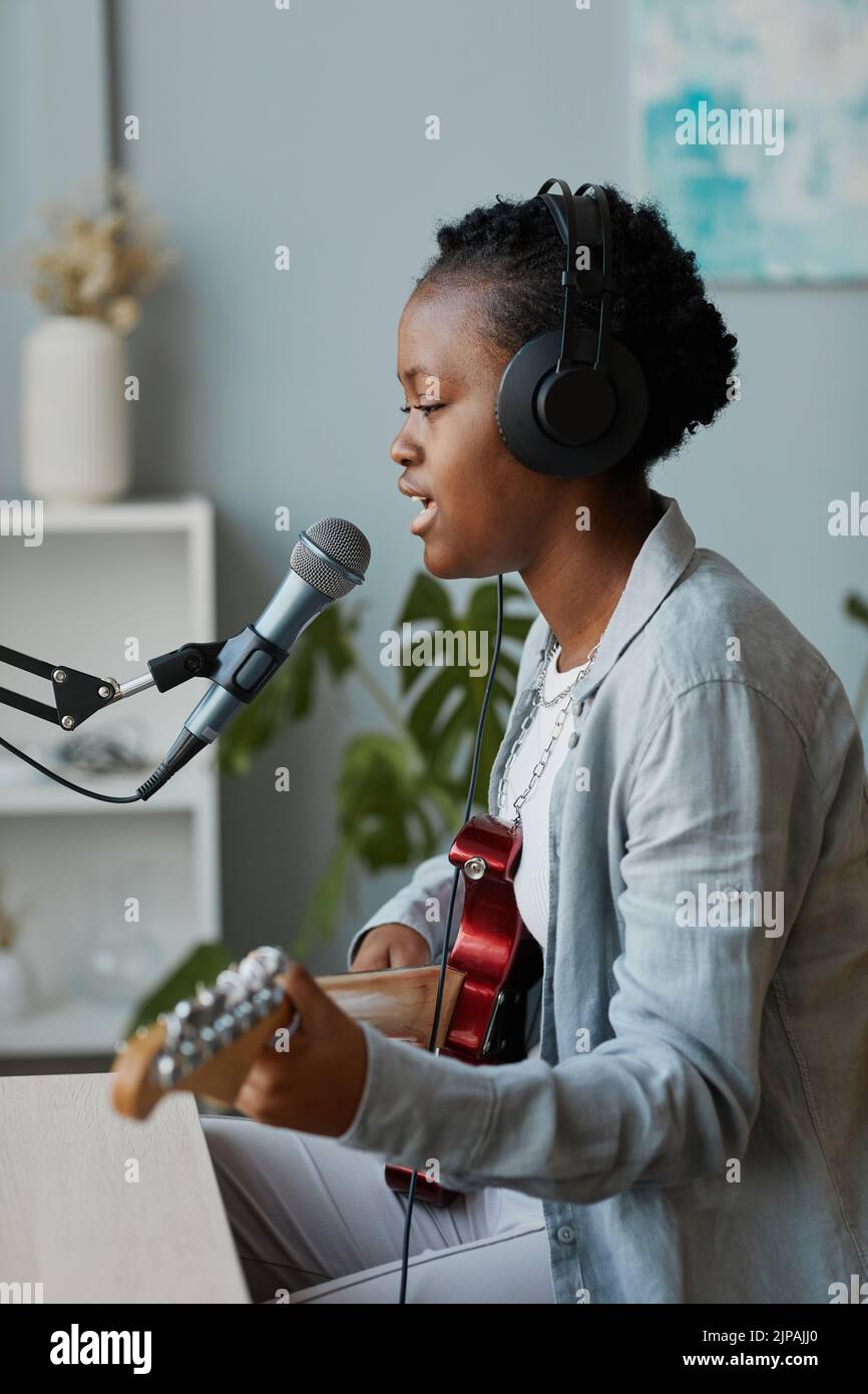 Vista lateral retrato de una joven mujer negra cantando al micrófono y tocando la guitarra en el estudio de grabación en casa Foto de stock