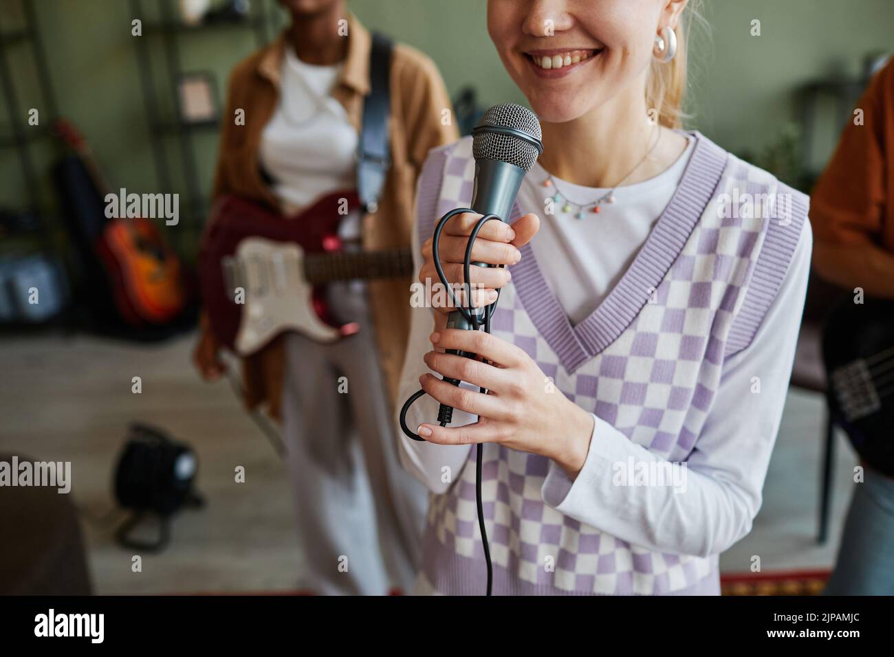 Recortada foto de una joven sonriente sosteniendo el micrófono mientras canta con la banda de música, espacio de copia Foto de stock