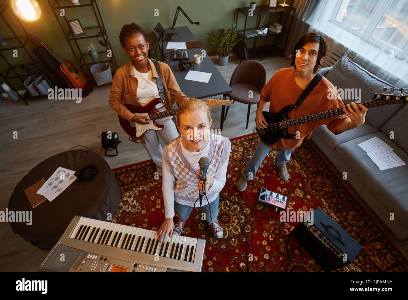 Retrato en ángulo alto de una banda de música que practica en un estudio en casa con enfoque en una joven rubia que sostiene el micrófono y mira hacia la cámara Foto de stock