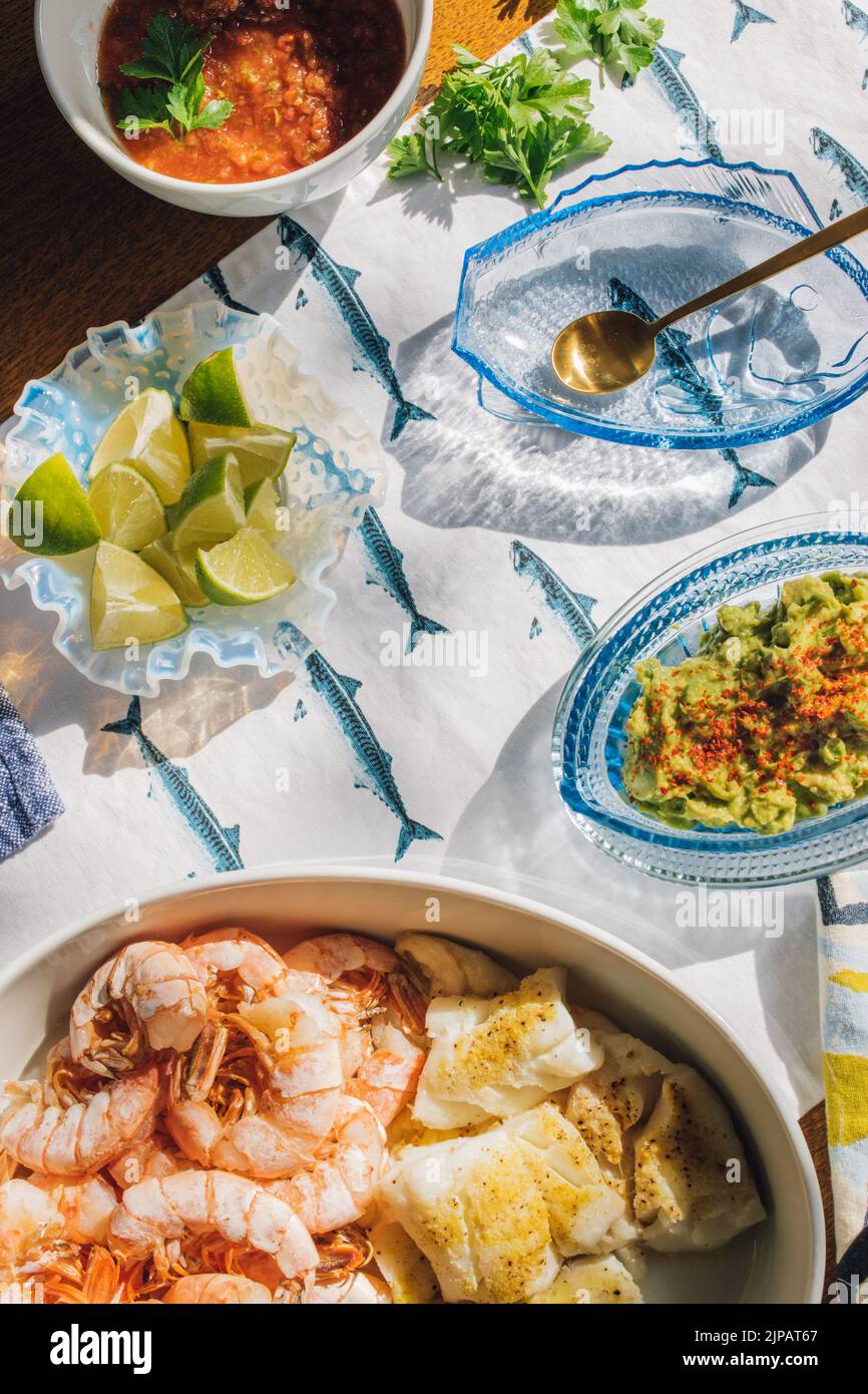 Mesa de cena de marisco de verano para hacer tacos de pescado, gambas, guacamole, lima, tortilla, Salsa sobre mantel de pescado azul y blanco al sol por Nicole Foto de stock