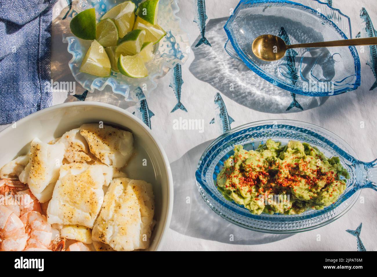 Mesa de cena de marisco de verano para hacer tacos de pescado, gambas, guacamole, lima, tortilla, salsa sobre mantel de pescado azul y blanco bajo el sol Foto de stock