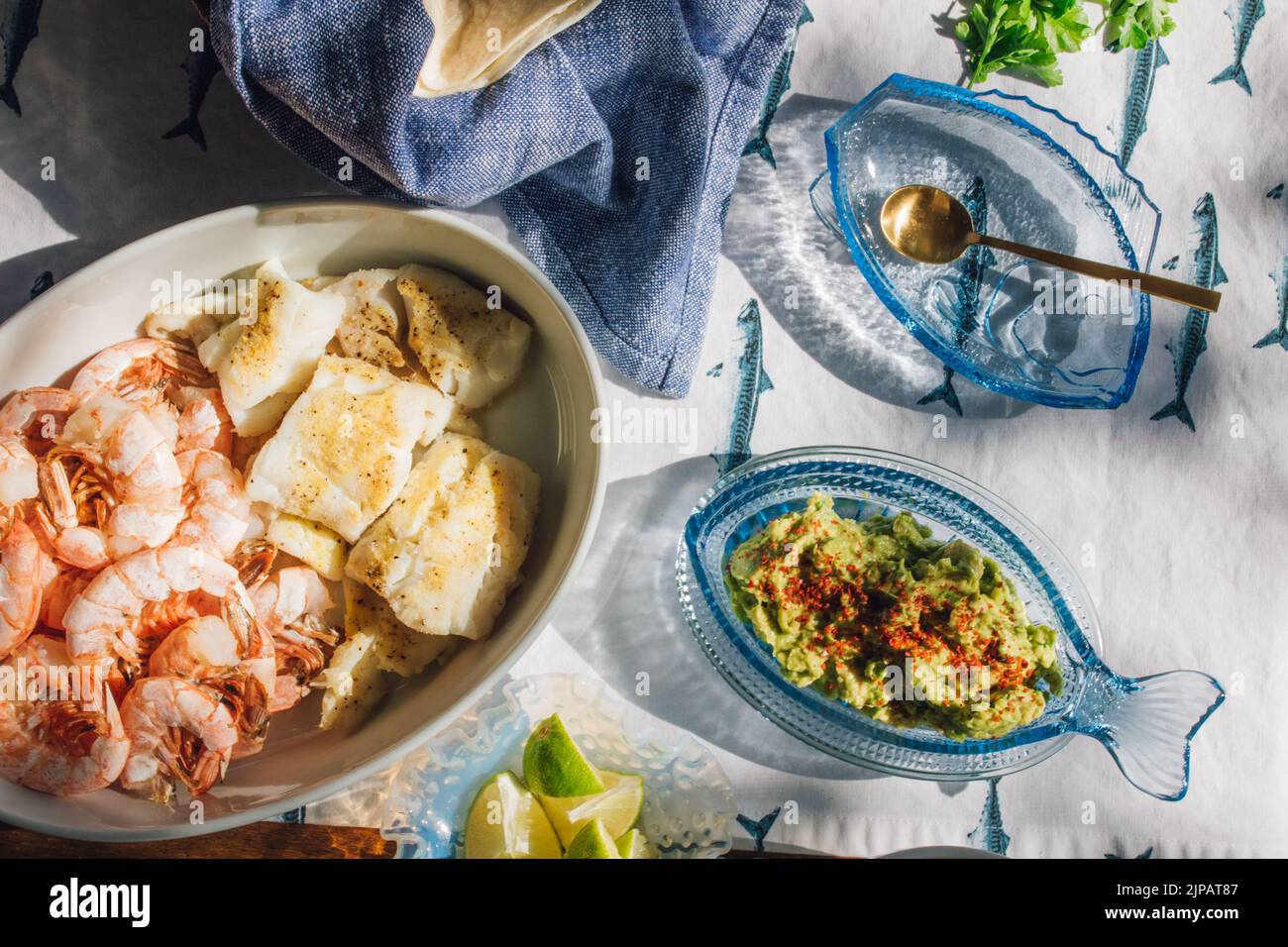 Mesa de cena de marisco de verano para hacer tacos de pescado, gambas, guacamole, lima, tortilla, salsa sobre mantel de pescado azul y blanco bajo el sol Foto de stock