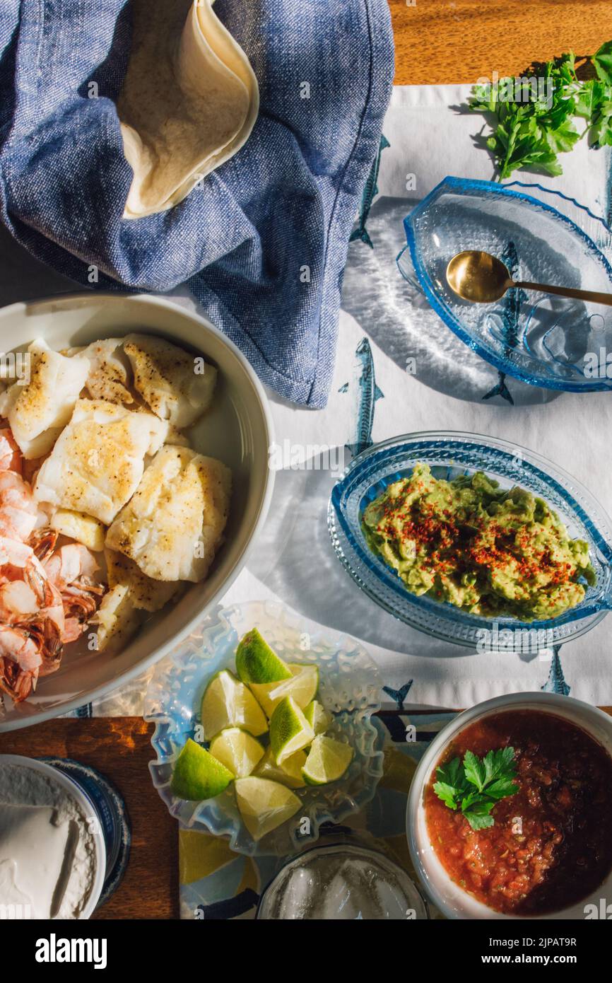Mesa de cena de marisco de verano para hacer tacos de pescado, gambas, guacamole, lima, tortilla, salsa sobre mantel de pescado azul y blanco bajo el sol Foto de stock
