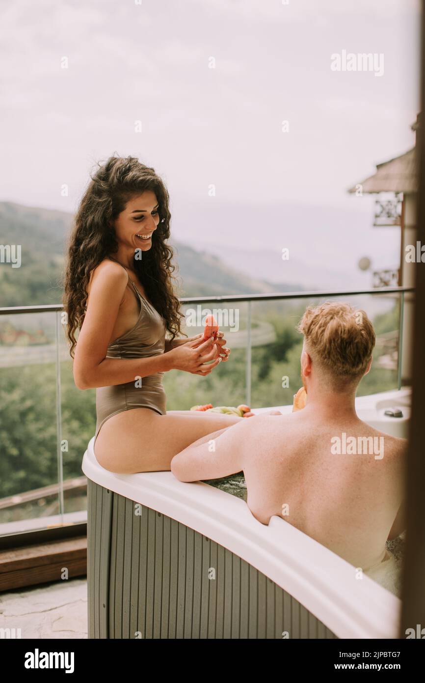 Atractiva pareja joven disfrutando en la bañera de hidromasaje al aire libre de vacaciones y comiendo fruta fresca Foto de stock
