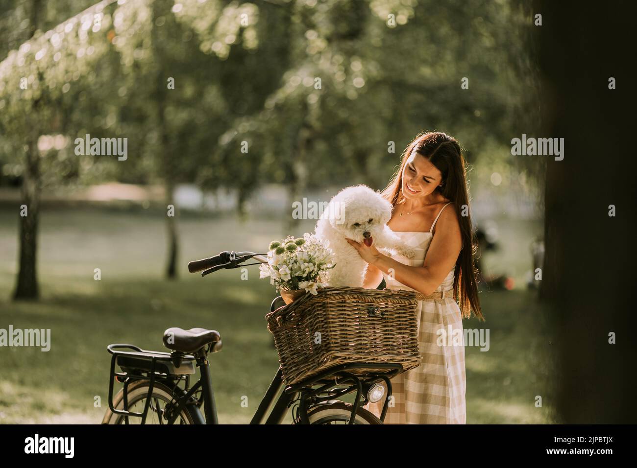 Una mujer joven con un perro blanco bichon en la cesta de la bicicleta eléctrica Foto de stock