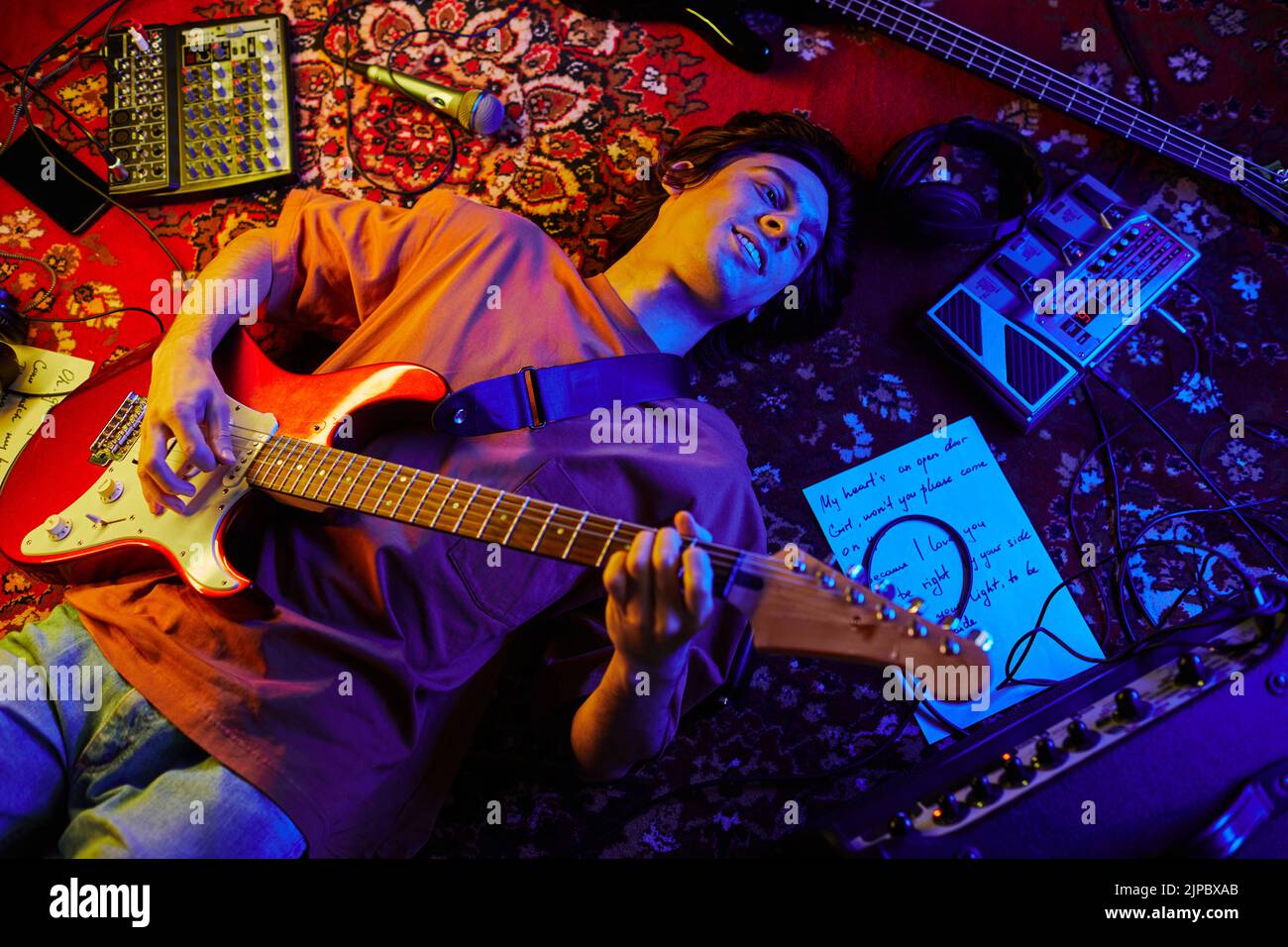 Imagen superior de un joven músico tocando la guitarra tumbado sobre una alfombra iluminada por la luz de neón Foto de stock