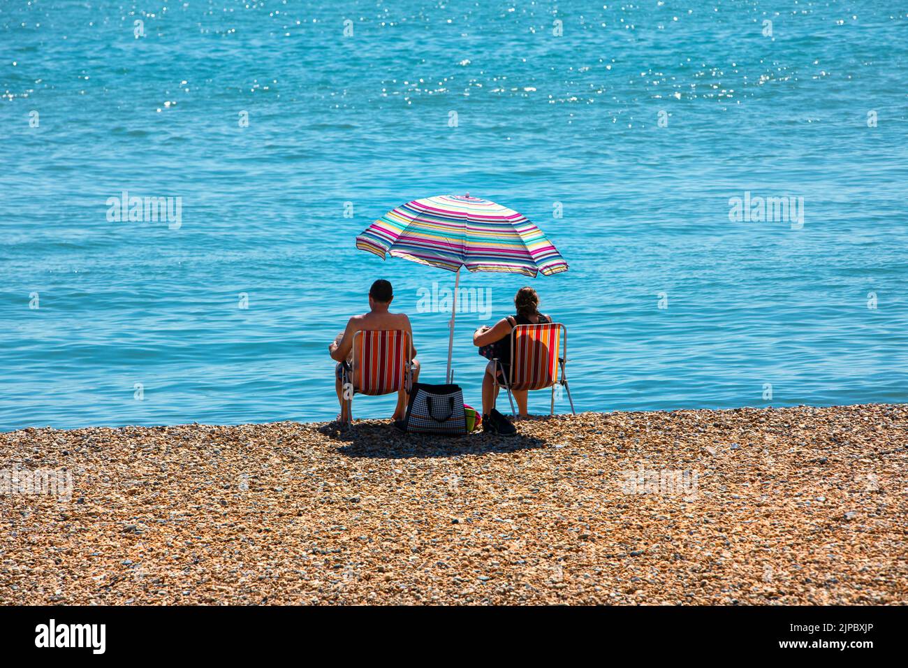 Una pareja en sillas de playa en una playa de guijarros junto a un mar azul y tranquilo. Foto de stock