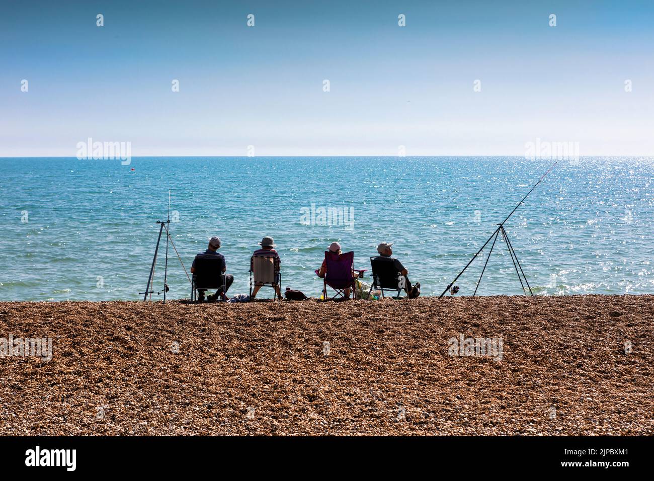 Cuatro pescadores sentados en una playa de guijarros junto a un mar azul en silueta Foto de stock