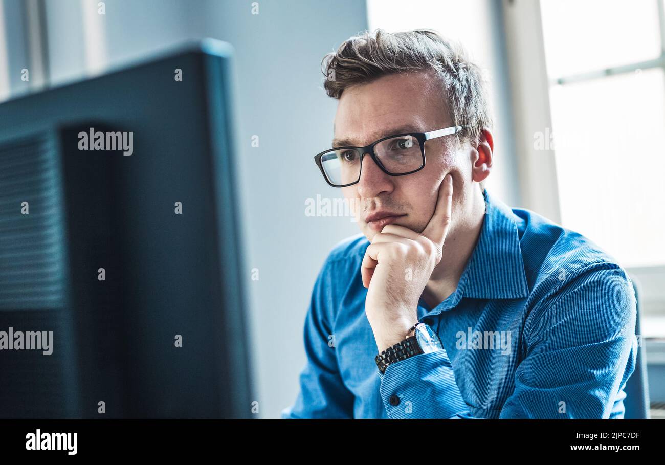 Guapo Joven hombre de negocios usando anteojos sentado en su mesa dentro de la oficina, mirando el informe en la pantalla de su computadora. Foto de stock