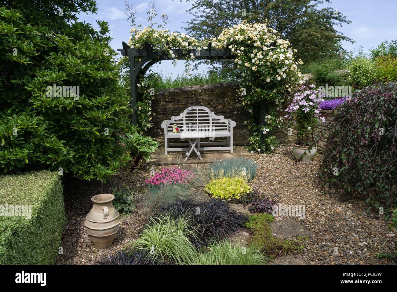 Un jardín inglés en verano, Gayton, Northamptonshire, Reino Unido; arbour, banco blanco y plantas que crecen en grava. Foto de stock