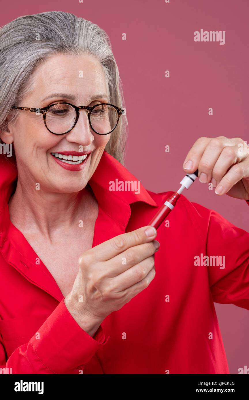 Mujer en camisa roja brillante sosteniendo brillo de labio y mirando contento Foto de stock