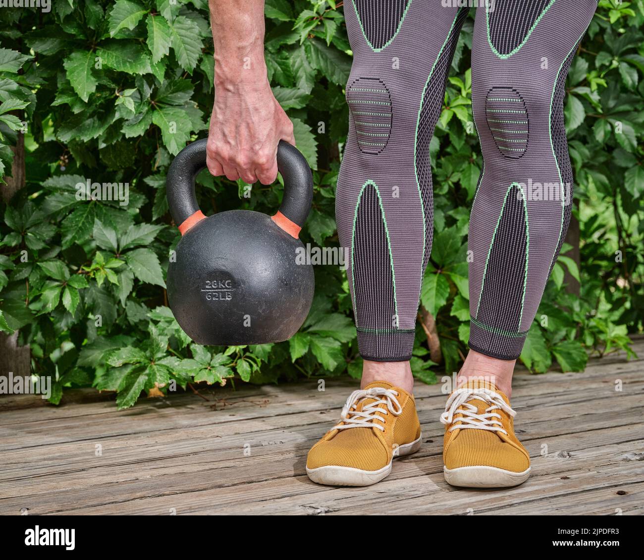 el hombre que usa refuerzos de compresión de rodilla y pantorrilla está haciendo ejercicio con una pesa de hierro en una cubierta de madera de patio trasero Foto de stock