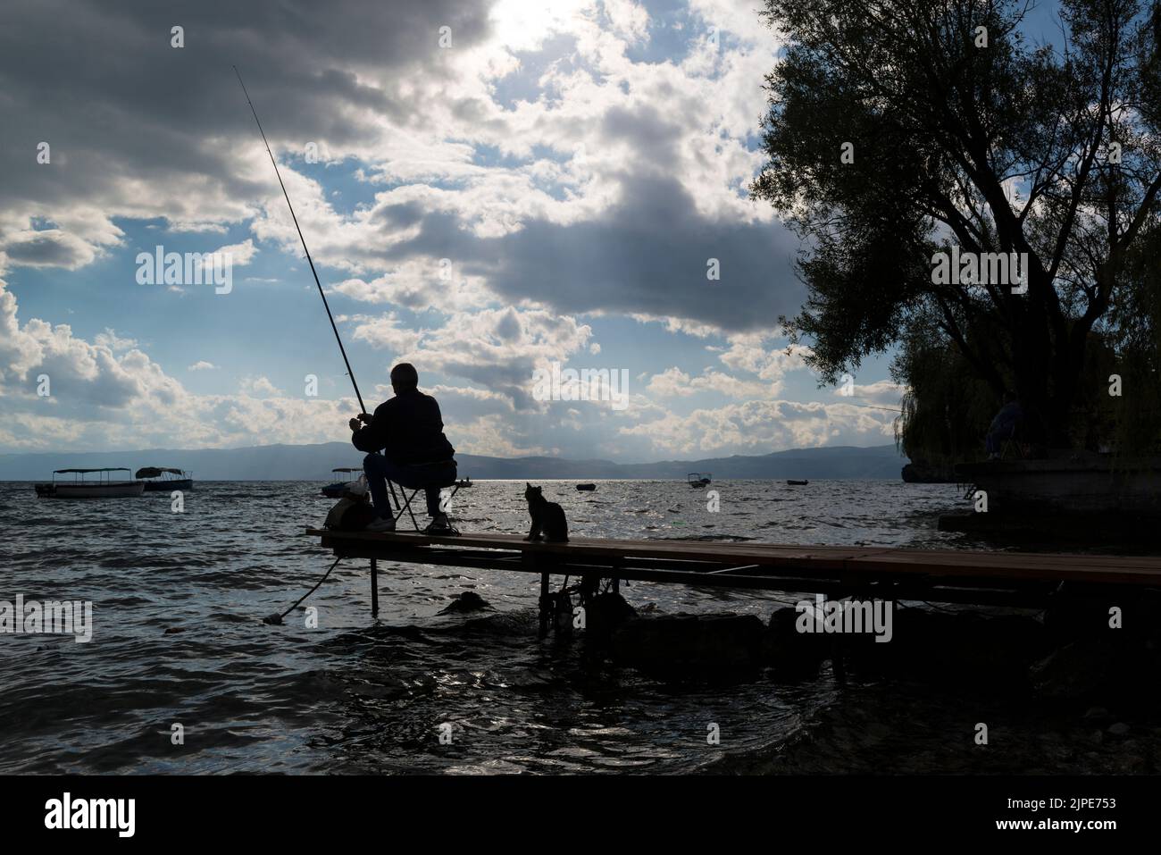 Silueta de un pescador deportivo y un gato oportunista detrás de él en un pequeño embarcadero en el lago Ohrid, en el norte de Macedonia. Foto de stock