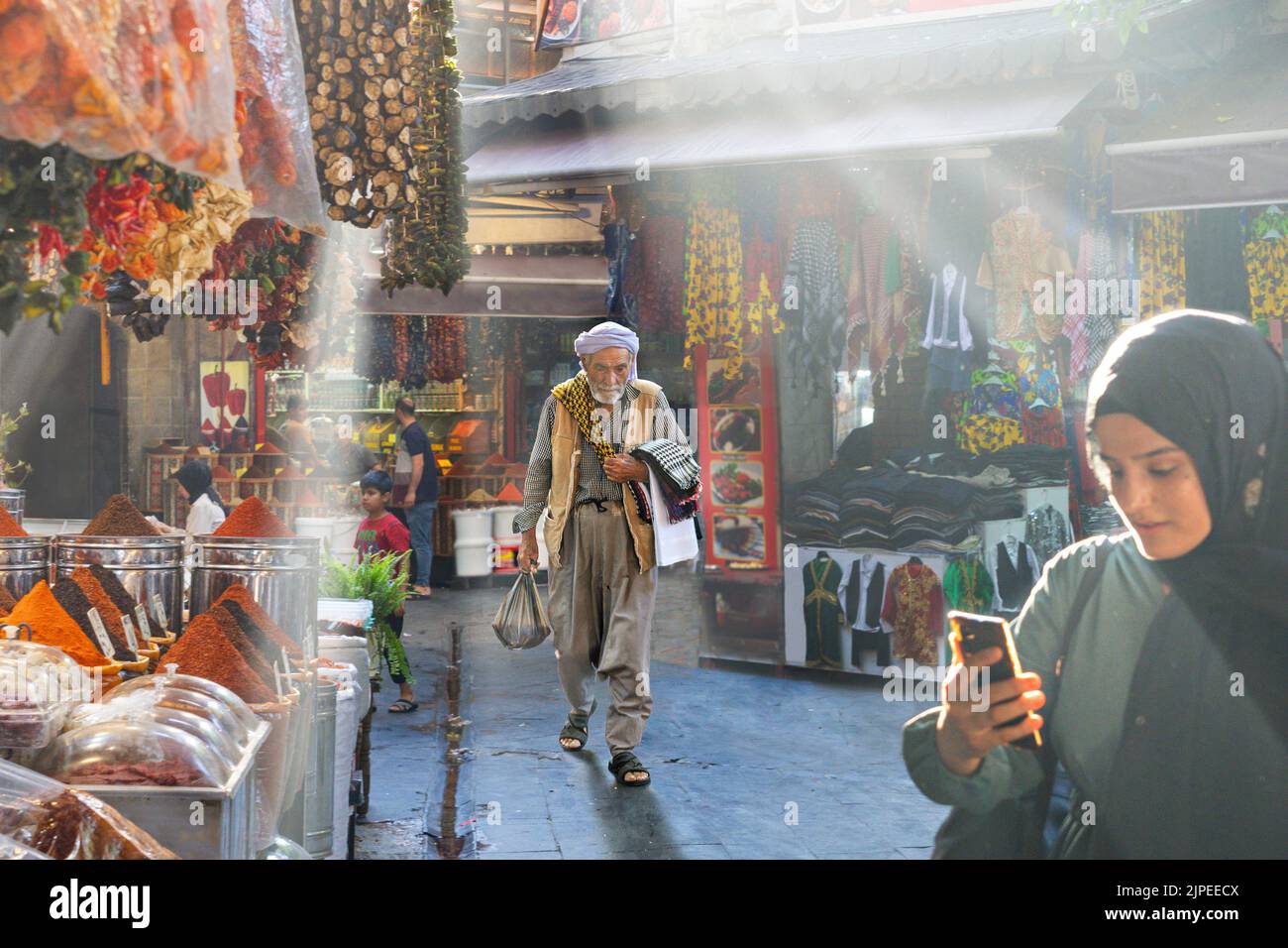 Mujer local revisando su teléfono y un hombre local con ropa tradicional caminando en el bazar a través de los rayos de luz y humo en Sanliurfa, Turquía Foto de stock