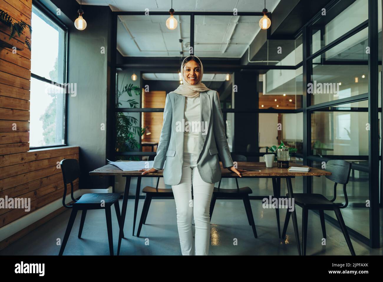 Feliz empresaria musulmana sonriendo a la cámara mientras estaba de pie en una sala de juntas de la oficina. Alegre empresaria usando un hijab en un moderno trabajo Foto de stock