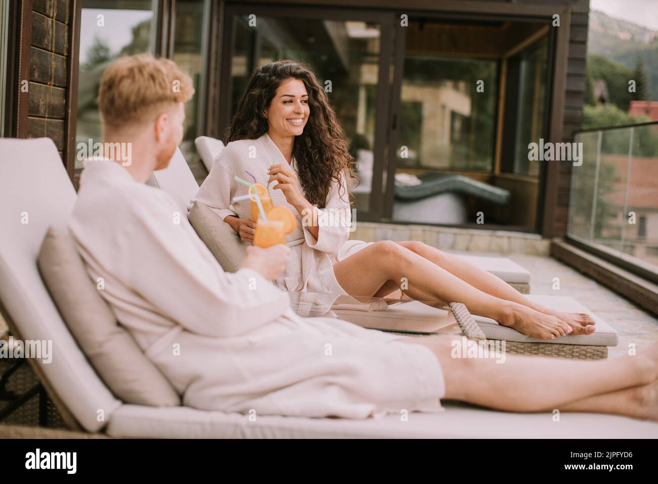 Una hermosa pareja joven que se relaja en las camas y bebe zumo de naranja fresco en la terraza al aire libre Foto de stock