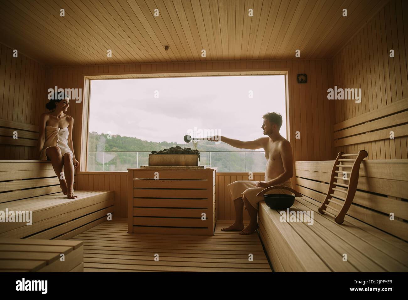 Hermosa joven pareja que se relaja en la sauna, mientras el hombre vertiendo agua sobre rocas calientes Foto de stock