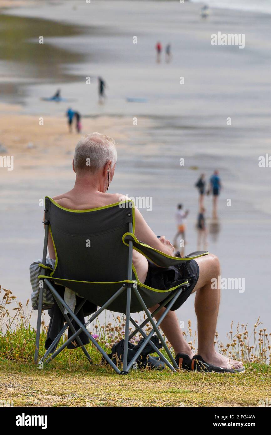 Un hombre maduro de vacaciones sentado en una silla plegable en la costa con vistas a la playa de Fistral en Newquay en Cornwall en el Reino Unido. Foto de stock