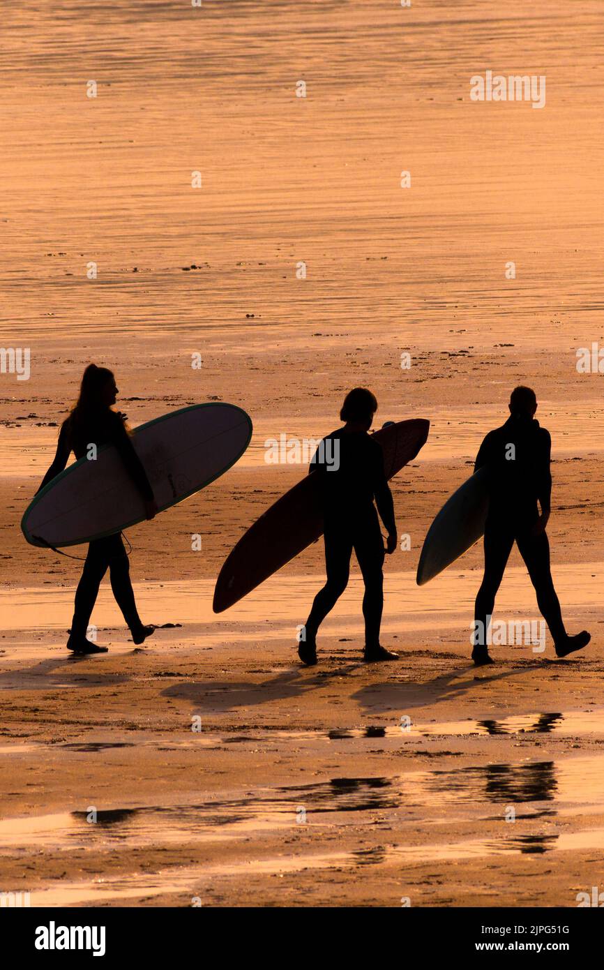 Un grupo de surfistas llevando sus tablas de surf y caminando por la silueta de la playa Fistral, representada por una intensa puesta de sol en Newquay, en Cornwall, Reino Unido. Foto de stock