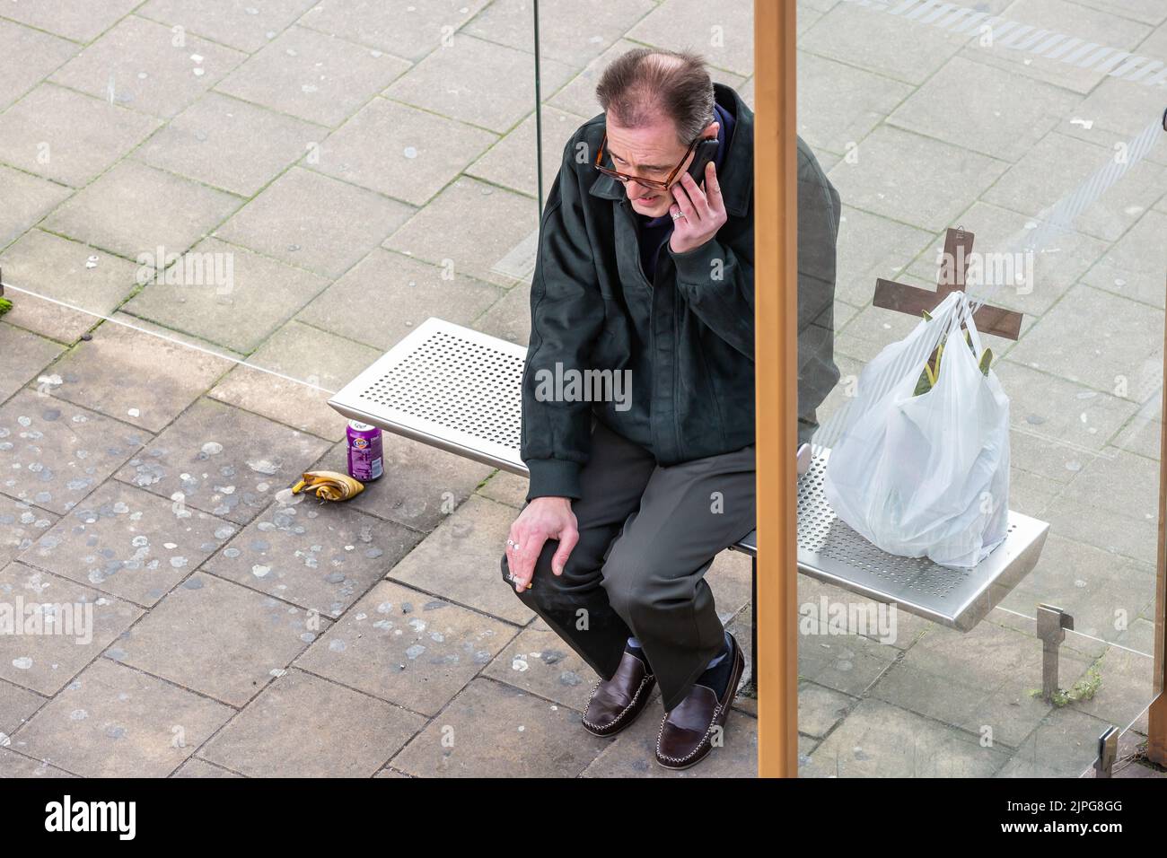 Hombre al teléfono, sentado en el banco de un refugio de autobuses, una bolsa de plástico que contiene una cruz a su lado. Foto de stock