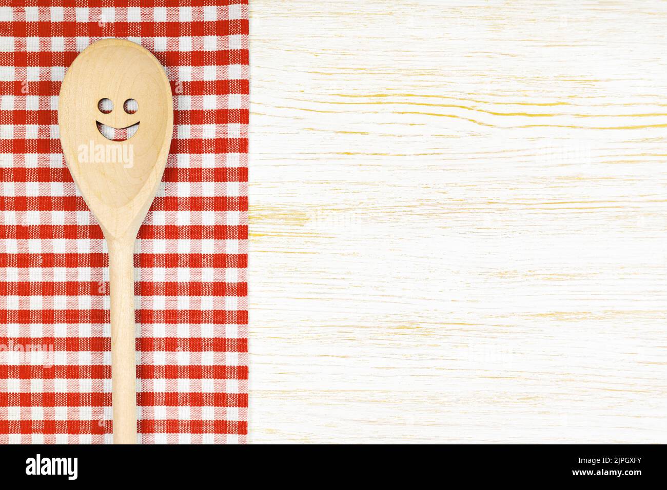 Cuchara de madera con sonrisa sobre mantel rojo a cuadros sobre superficie de madera blanca. Mockup para menú o receta, restaurante, sitio web con cocina. Cocina de alimentos Foto de stock