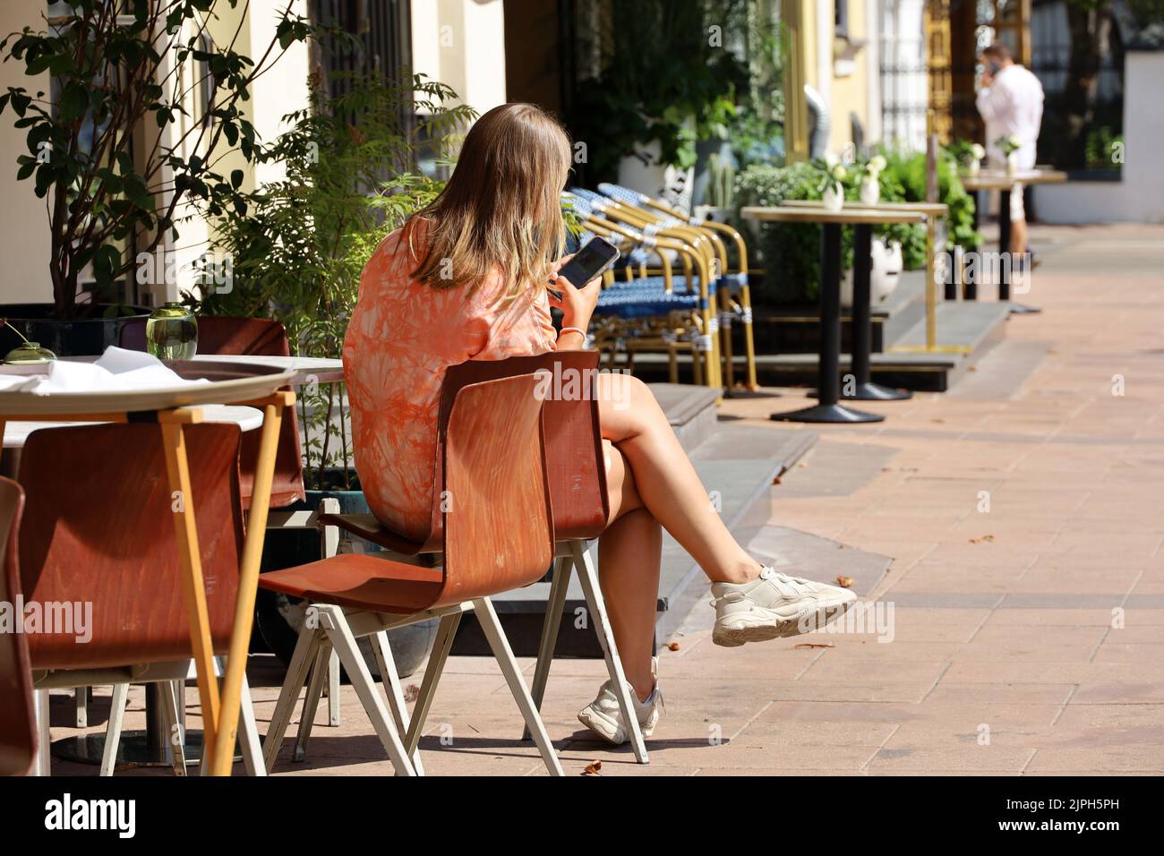 Chica en zapatillas de deporte sentada con un smartphone en una mesa en el café de la calle. Concepto de almuerzo solo, a la espera de una fecha o reunión de negocios, pausa para el café Foto de stock