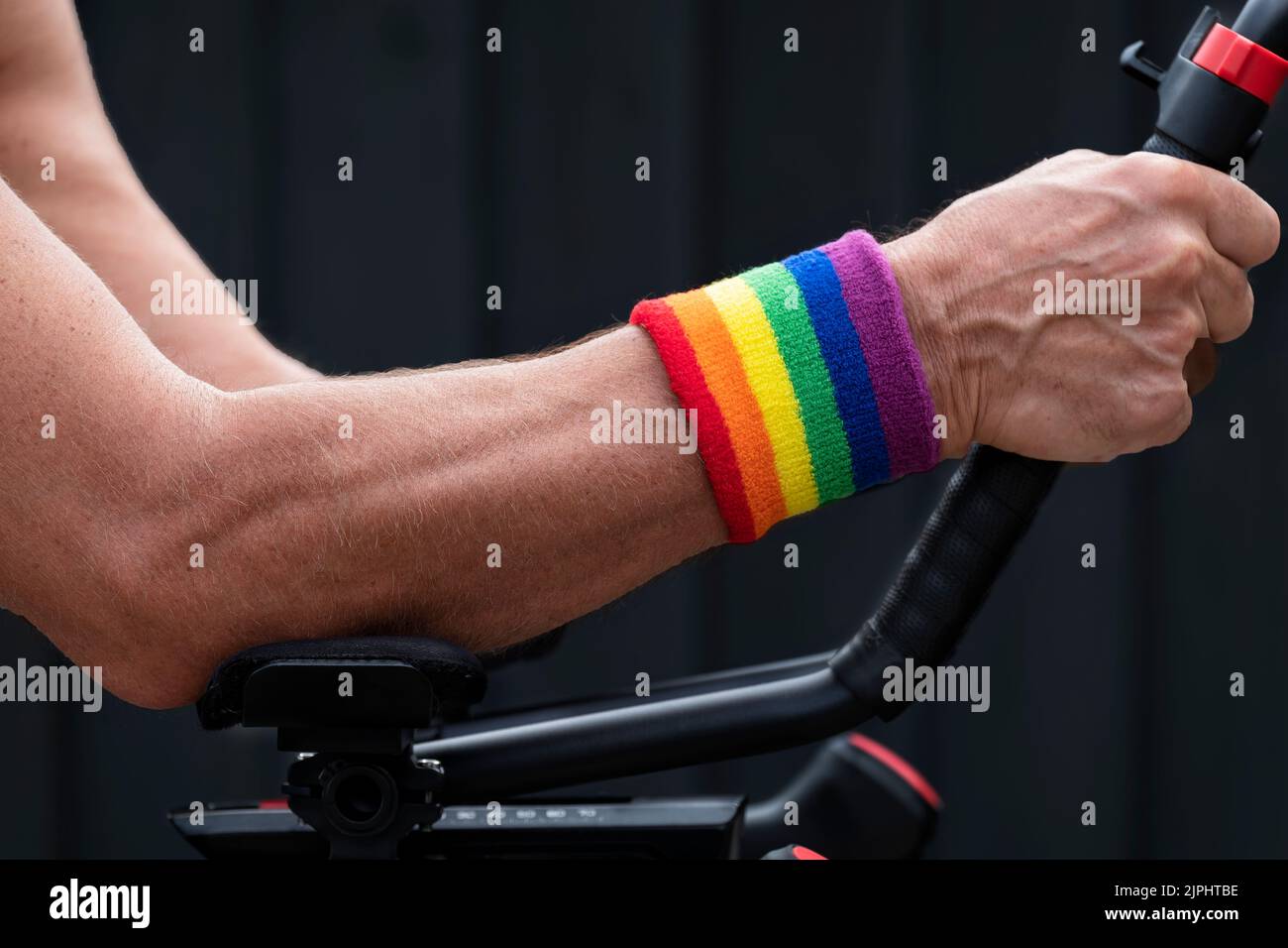 Un ciclista masculino que lleva un orgullo gay, una muñequera de tejido de rizo color arco iris. El ciclista se sostiene en las barras aerodinámicas de su bicicleta. Foto de stock