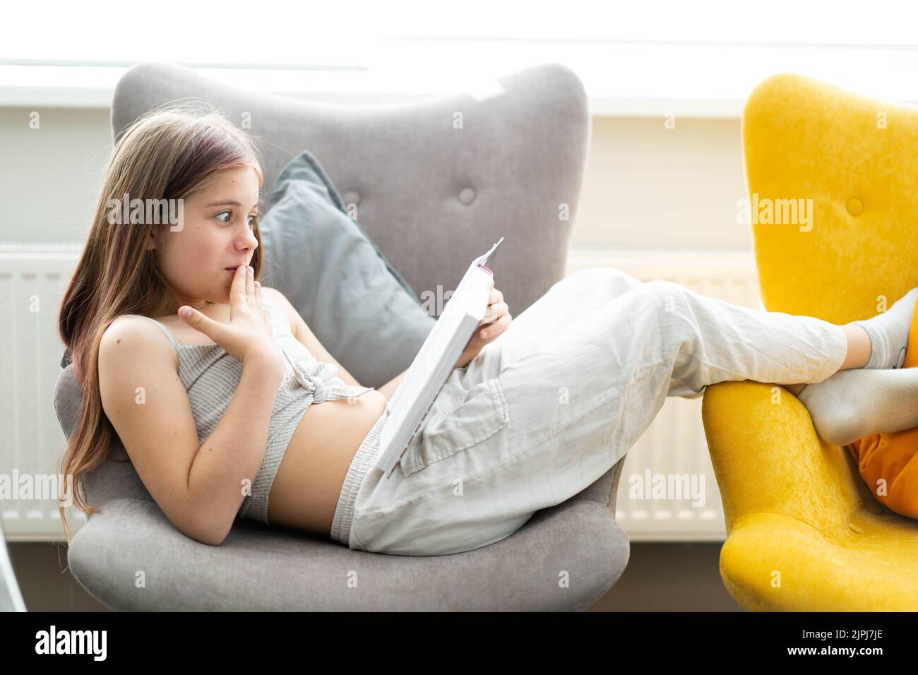 Retrato de una niña caucásica acostada en un sillón gris con un libro en las manos. La chica se ríe y se sorprende. El concepto de recreación en el hogar, aprendizaje, lectura. Foto de stock