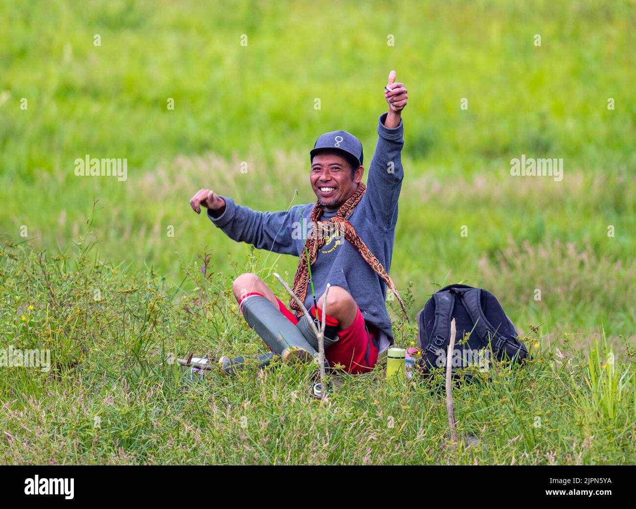 Un hombre indonesio alegre de mediana edad sentado en la hierba con el pulgar hacia arriba. Sulawesi, Indonesia. Foto de stock