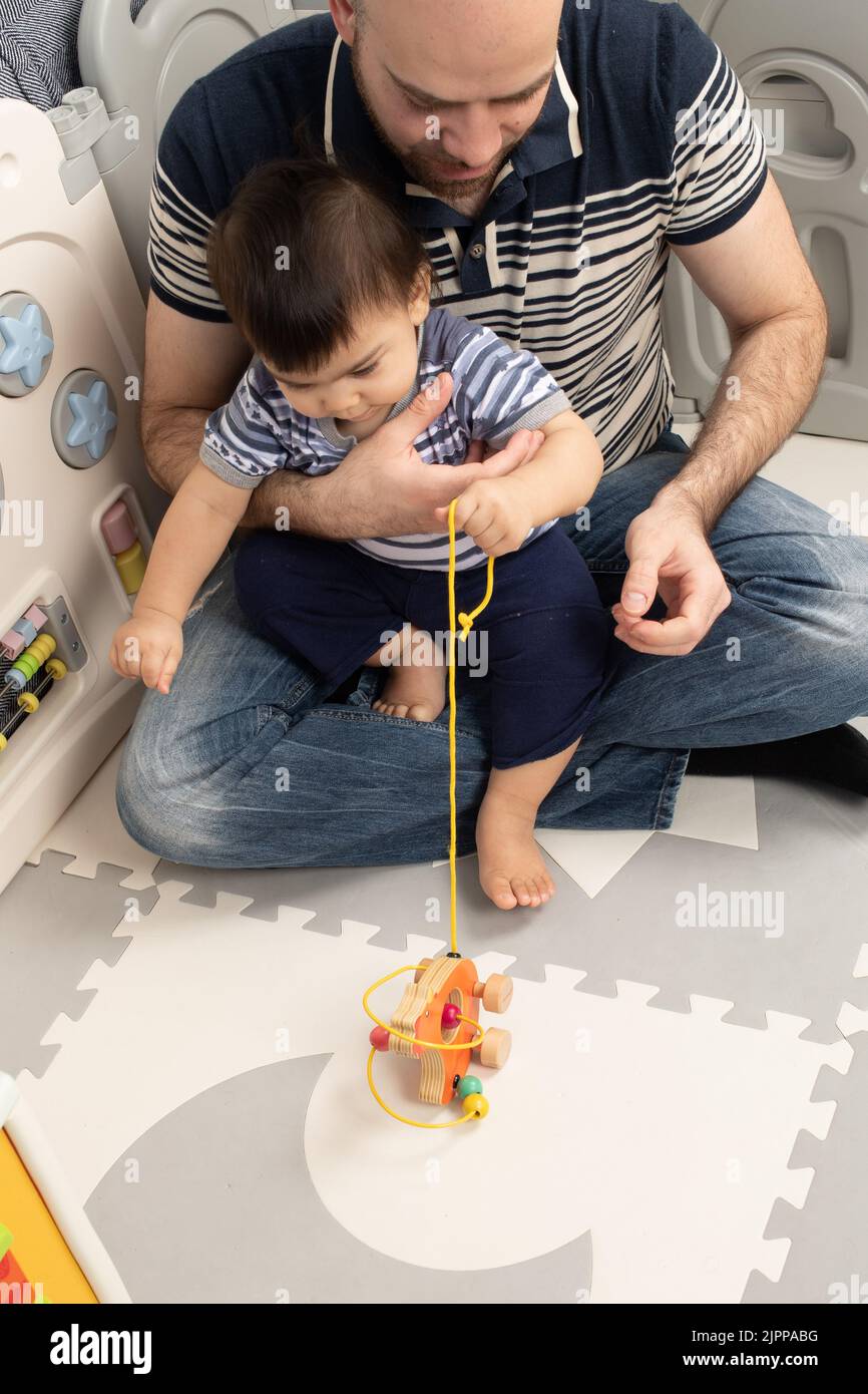 niño de 11 meses en casa con padre tirando de una cuerda para acercar un juguete con ruedas Foto de stock