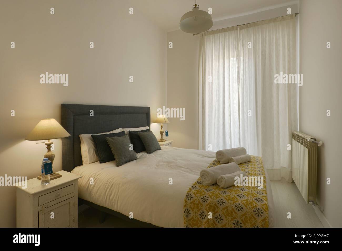 Dormitorio decorado en tonos suaves, cabecera tapizada en tela azul, cojines y lámparas a juego, cortinas blancas y toallas limpias Foto de stock