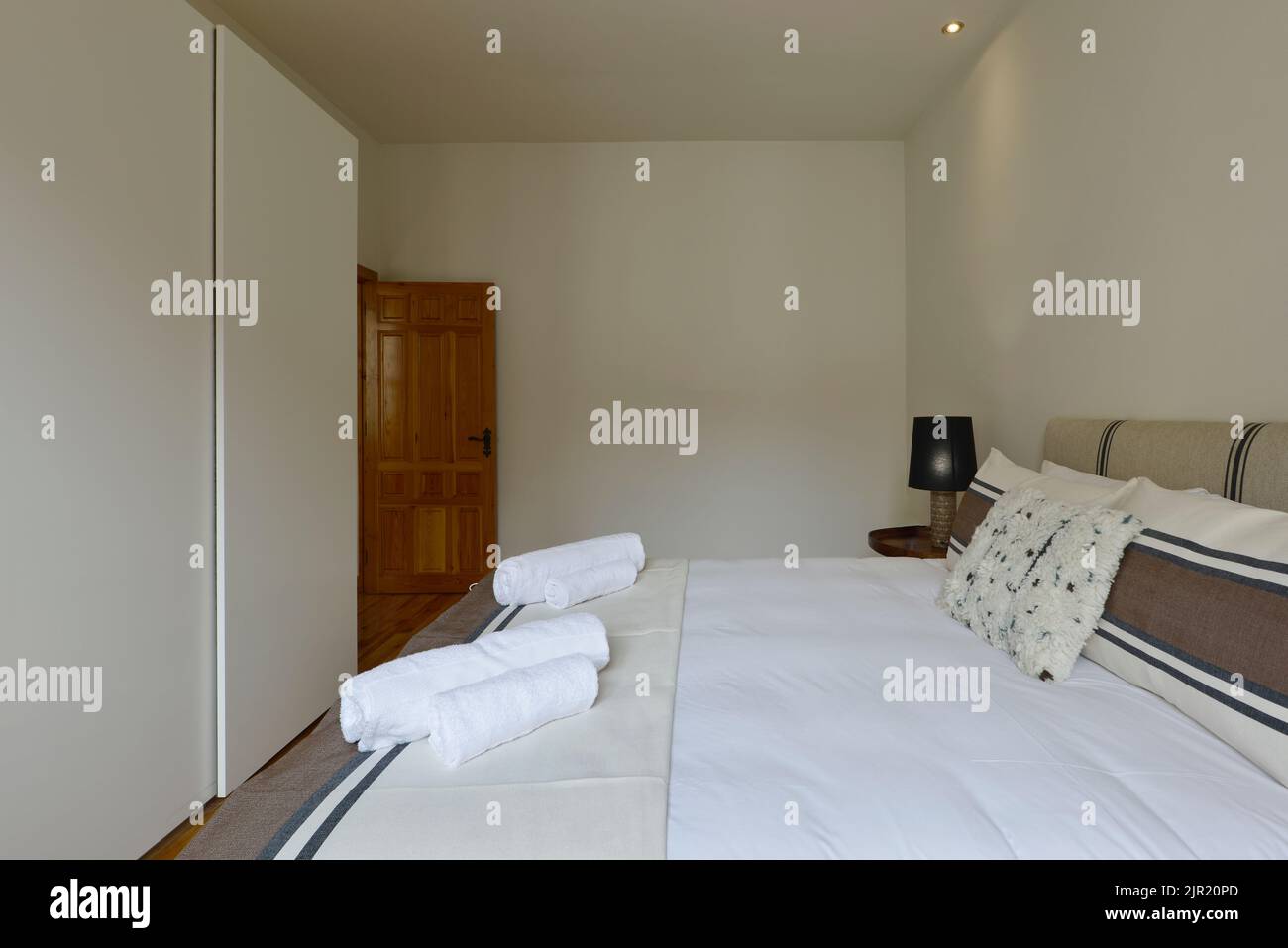 Dormitorio con cama doble, armario de puertas correderas de madera blanca, cojines y tapizados en tela y toallas limpias Foto de stock