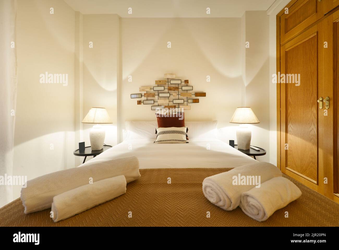 Dormitorio con cama doble con toallas enrolladas, armario con puertas de roble, cojines y 2 lámparas Foto de stock
