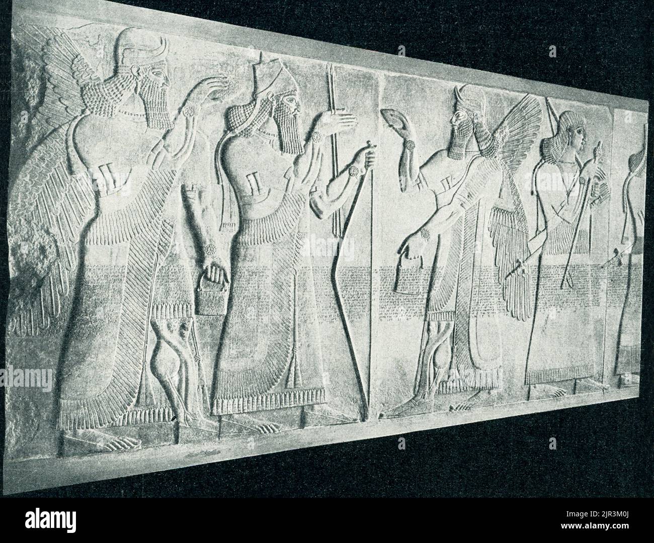El título de 1910 dice: 'Rey asirio entre genie - en el Museo Real de Berlín. La mítica figura guardiana asiria, llamada Genius, lleva un casco de dos cuernos y un vestido con flecos y lleva una daga de doble mango, todos los atributos de su naturaleza divina. Sus manos se mueven protectivamente hacia un árbol en flor, apenas visible en el borde derecho de la escultura. Este relieve, una vez pintado de forma brillante, es un fragmento del palacio del noroeste construido por el rey asirio Ashirnasirpal II (883-859 AEC) en su capital, Kalhu, ahora la ciudad iraquí de Nimrud. Docenas de enormes losas de piedra talladas con Foto de stock