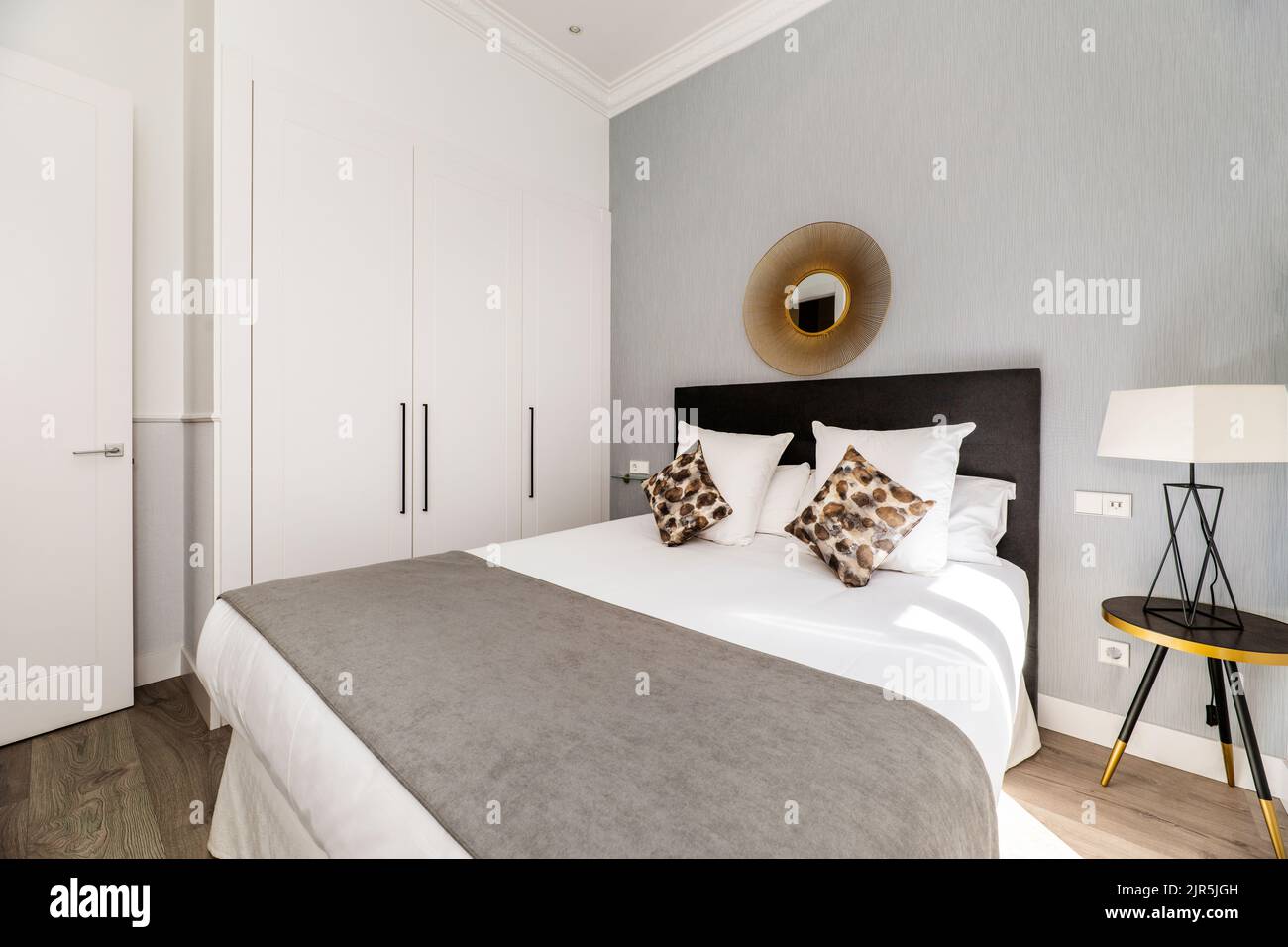 Dormitorio con cama doble con cabecera tapizada en tela marrón, cojines a juego y armario empotrado con puertas blancas Foto de stock