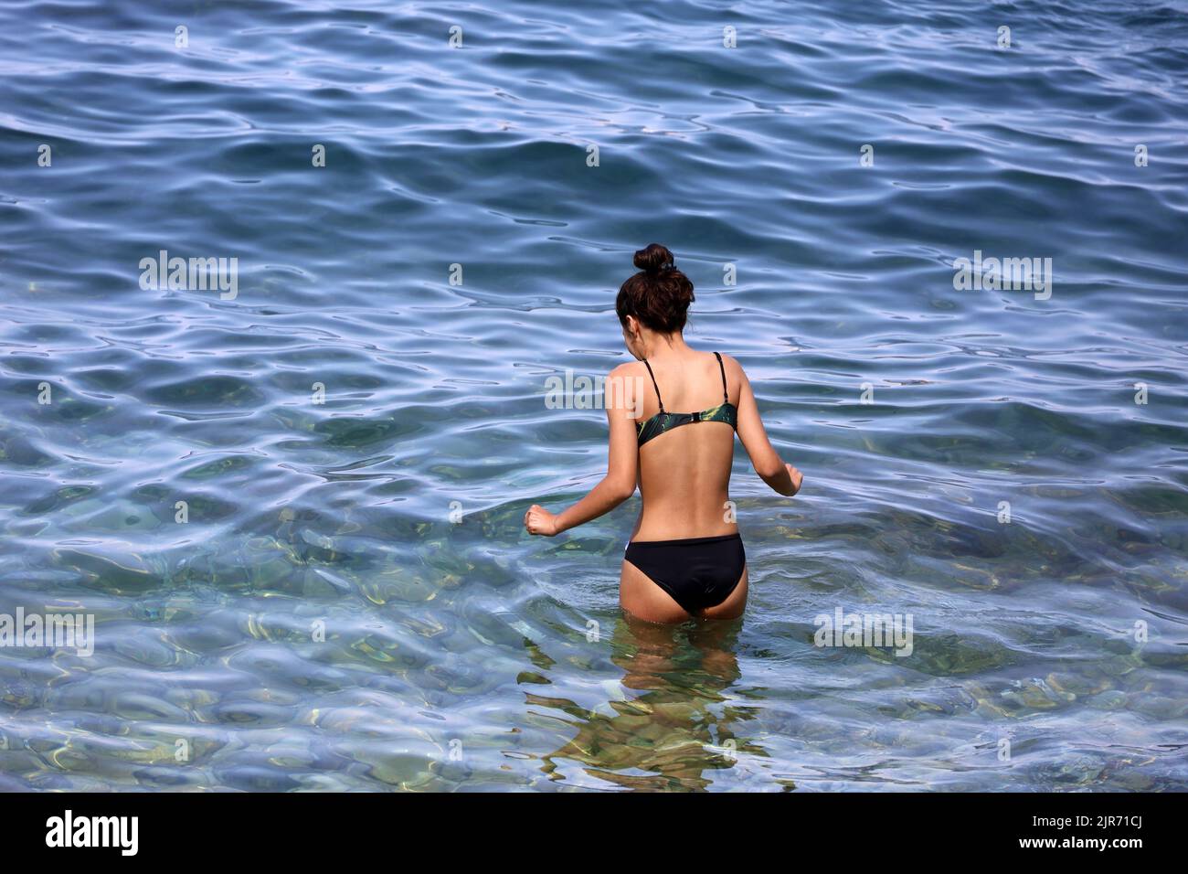 Chica en traje de baño negro va a nadar en el agua del mar. Vacaciones en una playa de guijarros de complejo de verano Foto de stock